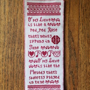 May include: A white cross-stitch embroidery with red thread, featuring a poem about love. The poem reads: "O My Luve is like a red, red Rose that's newly spruns in June. O My Luve is like the Melody that's sweetly played in tune."