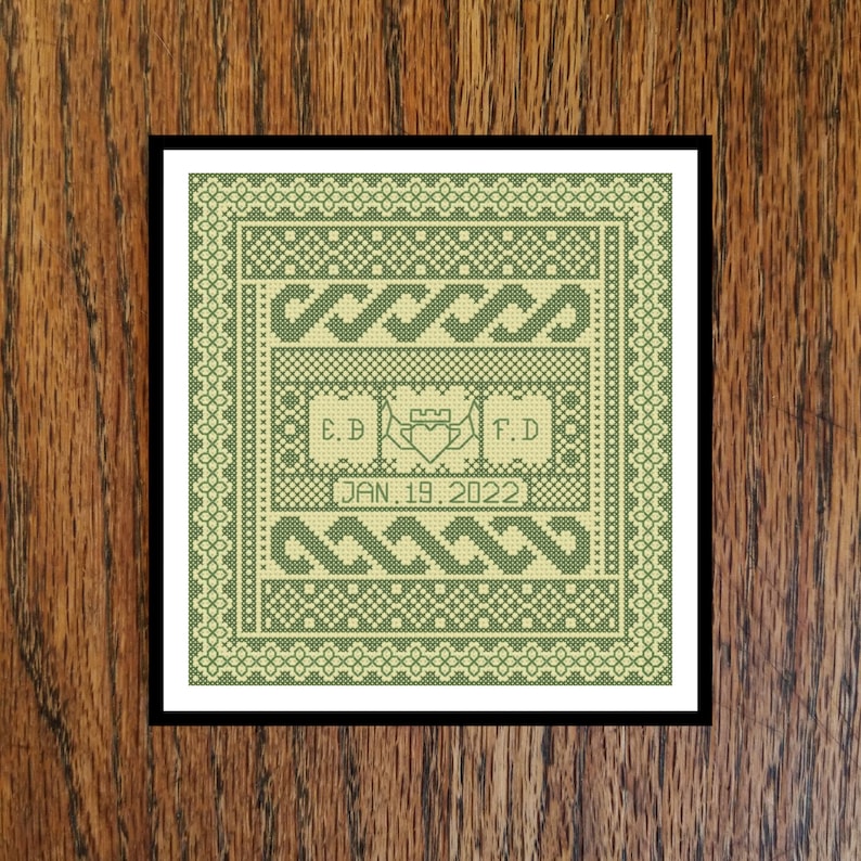 May include: A framed cross-stitch sampler in shades of green and cream. The design includes intertwined patterns, initials "E.D" and "F.D", a heart and crown emblem, and the date "JAN. 19, 2022".