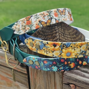 May include: A stack of three dog collars with floral patterns. The top collar has a white background with orange butterflies and flowers. The middle collar has yellow sunflowers, and the bottom collar has a dark blue floral design. Each collar has a green leather strap and gold hardware.