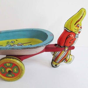 May include: Vintage tin toy wheelbarrow with a yellow and red rabbit figure. The wheelbarrow has a blue basin with pink and red accents, a yellow interior, and a red frame. The wheel is yellow with green and red details.