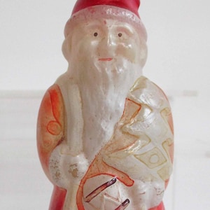 May include: A vintage Christmas light bulb shaped like Santa Claus. The bulb has a red hat and suit, with a white beard and trim. The design includes a sack and a small figure. The bulb is mounted on a white base.