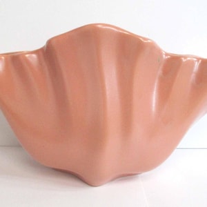 May include: A salmon-coloured ceramic bowl with a wide, flared opening and a scalloped, shell-like silhouette. The surface features a smooth, glossy finish with vertical ridges that radiate from the base towards the undulating, curved rim.