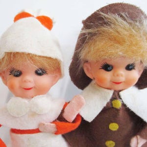 Peut inclure: Deux figurines de poupées vintage. L'une porte un chapeau blanc avec des accents orange et une tenue blanche avec des manches orange. L'autre porte un chapeau marron avec une garniture en fausse fourrure et un manteau marron avec des boutons jaunes. Les deux poupées ont les cheveux blonds et les joues roses.