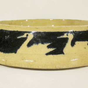 Puede incluir: Un cuenco de cerámica de color crema con un borde negro y un diseño de pájaros estilizados en negro. El cuenco está ligeramente acampanado en el borde.