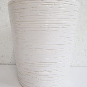 Puede incluir: Un jarrón alto de cerámica blanquecina con un patrón texturizado de líneas horizontales. El jarrón tiene forma cilíndrica y un acabado brillante, adecuado para la decoración del hogar.