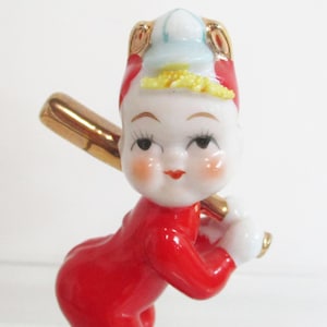 May include: A ceramic figurine of a character wearing a red outfit and a white cap with gold accents. The figure has a pale face with rosy cheeks, red lips, and dark eyelashes, and is holding a shiny gold baseball bat across its shoulders.