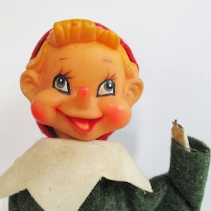 Puede incluir: Un muñeco de elfo vintage con un gorro rojo, un cuello de piel blanca y un traje de fieltro verde. El elfo tiene una cara pintada con mejillas sonrosadas y una amplia sonrisa.