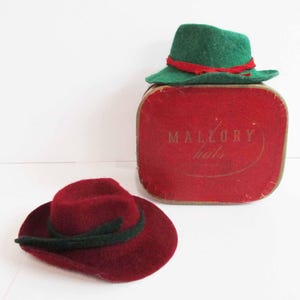 May include: Two felt hats, one green with a red band and one burgundy with a green band, sit near a red hat box. The hat box has the text "MALLORY hats" printed on it.
