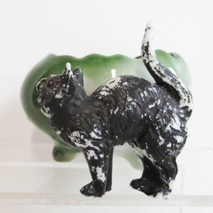 May include: A black cat figurine with white accents stands in front of a green ceramic planter. The cat has a textured appearance, with its tail arched upwards. The planter has a scalloped edge.