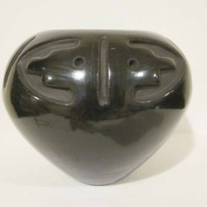 Op de afbeelding: Zwarte keramische pot met een gladde, glanzende afwerking. De pot heeft een ronde vorm met twee gestileerde gezichten die in de voorkant zijn gegraveerd.