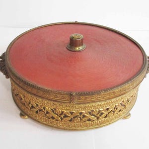 Pode incluir: Uma caixa de joias redonda e antiga com tampa vermelha e ornamentação em metal dourado. A caixa apresenta um padrão floral decorativo ao redor da base e um pequeno botão na tampa. Estética vintage.