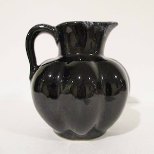 Puede incluir: Jarra de cerámica negra brillante con cuerpo redondeado y asa curva. La jarra tiene un borde festoneado y una forma bulbosa, que refleja la luz. El color oscuro y el diseño sencillo sugieren un estilo vintage o rústico.