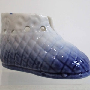 Puede incluir: Un zapato de bebé de cerámica azul y blanco con un patrón tejido y agujeros. El zapato está esmaltado y tiene un color azul descolorido.