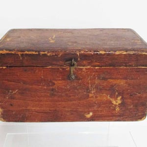 Puede incluir: Una caja rectangular de madera con un acabado marrón oscuro y signos visibles de desgaste, incluyendo arañazos y bordes desgastados. La caja cuenta con una tapa abatible asegurada por un pequeño mecanismo de cierre metálico centrado en el frente.