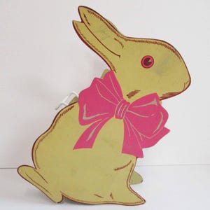 Puede incluir: Decoración vintage de conejito de Pascua. El conejito es amarillo claro con un lazo rosa y un contorno marrón. El conejito tiene un ojo rosa y está sentado erguido. La decoración es de cartón.