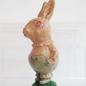May include: A standing rabbit figurine with a tan upper body, light green pants, and a dark green circular base. The rabbit has painted red eyes and a textured surface with visible signs of wear, including small dark spots across the paint.
