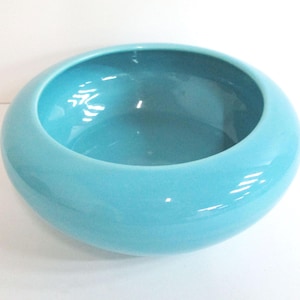 May include: A round, turquoise ceramic bowl with a glossy finish. The bowl has a wide, shallow design and a smooth, curved rim. The interior of the bowl reflects light, creating highlights. This decorative bowl is suitable for various uses.