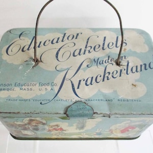 May include: Vintage blue tin box with a metal handle, featuring the text "Educator Cakelets Made in Krackerland." The box has a light blue background with cloud illustrations and the text "Johnson Educator Food Co. Cambridge, Mass., U.S.A."