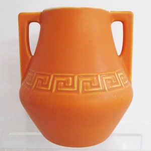 May include: Orange ceramic vase with two handles. The vase features a geometric pattern around the middle. The vase has a wide body and a narrow neck. The handles are rectangular and attached to the sides of the vase.