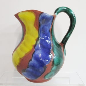 May include: A colorful ceramic pitcher with a curved handle. The pitcher features a design of vertical stripes in yellow, blue, and green against a reddish-brown background. The handle is a glossy green.