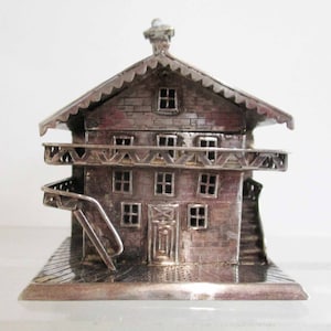 May include: A silver miniature house with a gabled roof, a balcony, and a staircase. The house has a detailed exterior with windows and a door.