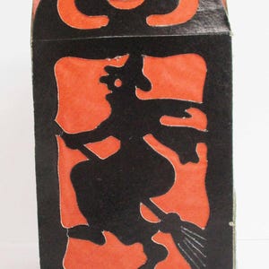 May include: A vintage Halloween decoration featuring a black silhouette of a witch on a broomstick against an orange background. The top has a smiling jack-o'-lantern face. The box is made of cardboard and is a festive holiday item.