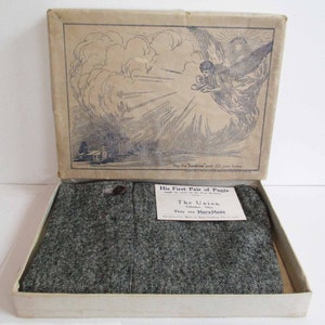 May include: An open box with a pair of gray pants inside. The box is decorated with a drawing of an angel holding a baby. The label on the box reads "His First Pair of Pants" and "The Union, Columbus, Ohio. They are Marx Made."