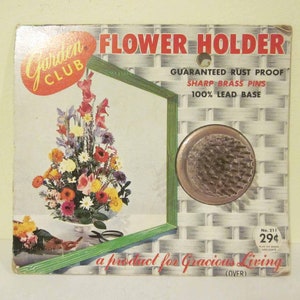 Op de afbeelding: Een vintage bloemenhouder met een roestvrij loodbasis en scherpe messing pinnen. De verpakking toont een kleurrijk bloemstuk en de tekst "Garden Club Flower Holder, Guaranteed Rust Proof, Sharp Brass Pins, 100% Lead Base, No. 211, 29¢, a product for Gracious Living (OVER)"