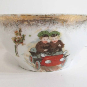 Puede incluir: Un tazón de porcelana blanca con un borde dorado y una ilustración colorida de dos niños en un coche rojo. Los niños llevan sombreros verdes y abrigos de piel. El tazón tiene un aspecto vintage.