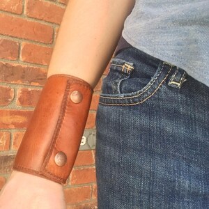 May include: A brown leather wrist cuff with two copper-colored button closures. The cuff is worn on the wrist and is made of stitched leather. The background is a red brick wall and blue jeans.