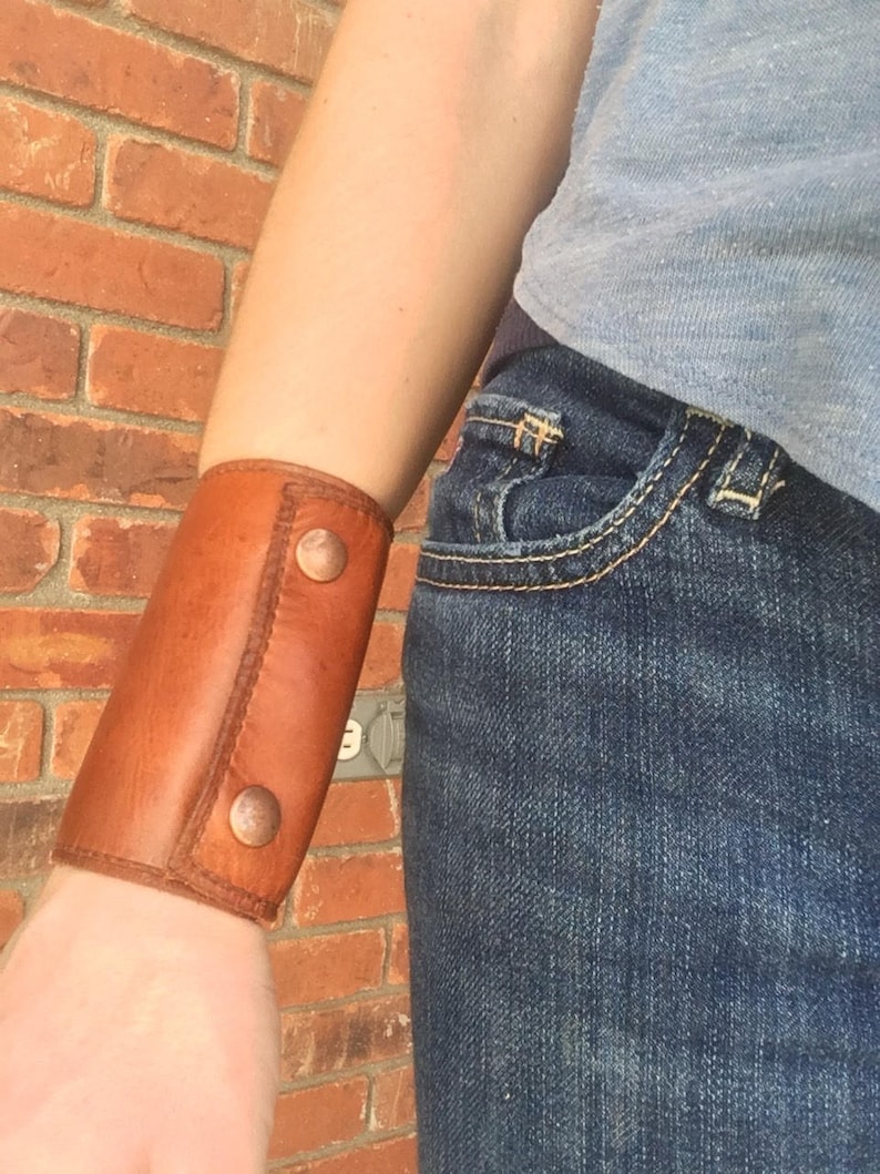 May include: A brown leather wrist cuff with two copper-colored button closures. The cuff is worn on the wrist and is made of stitched leather. The background is a red brick wall and blue jeans.