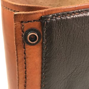 May include: Close-up of a bi-color leather wallet. The wallet features a combination of brown and black leather, with visible stitching and a metal snap closure. The leather appears to be textured, and the design is simple and classic.