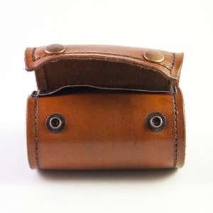 May include: A small, open, brown leather case with a rounded rectangular shape. The case has a flap secured by two copper-colored rivets. Two additional black rivets are visible on the body of the case. The leather has a glossy finish.