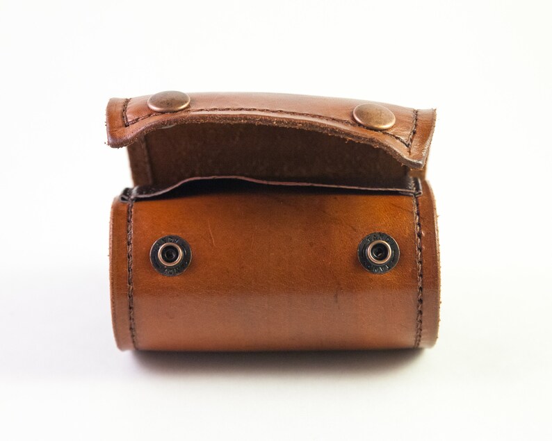 May include: A small, open, brown leather case with a rounded rectangular shape. The case has a flap secured by two copper-colored rivets. Two additional black rivets are visible on the body of the case. The leather has a glossy finish.