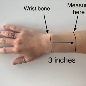 May include: A close-up of a person's wrist with a black line drawn around it. The text "Measure here" is written above the line. An arrow points to the right with the text "3 inches" below it. The text "Wrist bone" is written above the wrist with an arrow pointing down.