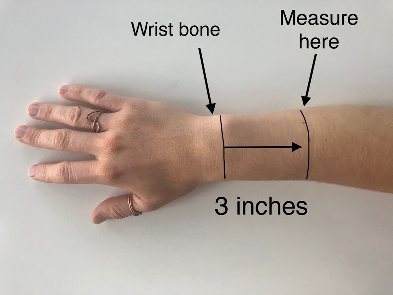 May include: A close-up of a person's wrist with a black line drawn around it. The text "Measure here" is written above the line. An arrow points to the right with the text "3 inches" below it. The text "Wrist bone" is written above the wrist with an arrow pointing down.
