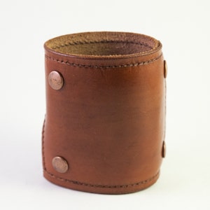 May include: A brown leather cuff bracelet with copper-colored rivets. The cuff is cylindrical and appears to be made of a single piece of leather, with a stitched seam. The rivets are evenly spaced around the cuff.