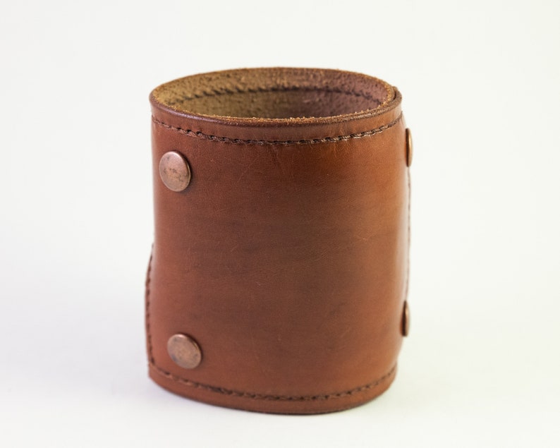 May include: A brown leather cuff bracelet with copper-colored rivets. The cuff is cylindrical and appears to be made of a single piece of leather, with a stitched seam. The rivets are evenly spaced around the cuff.