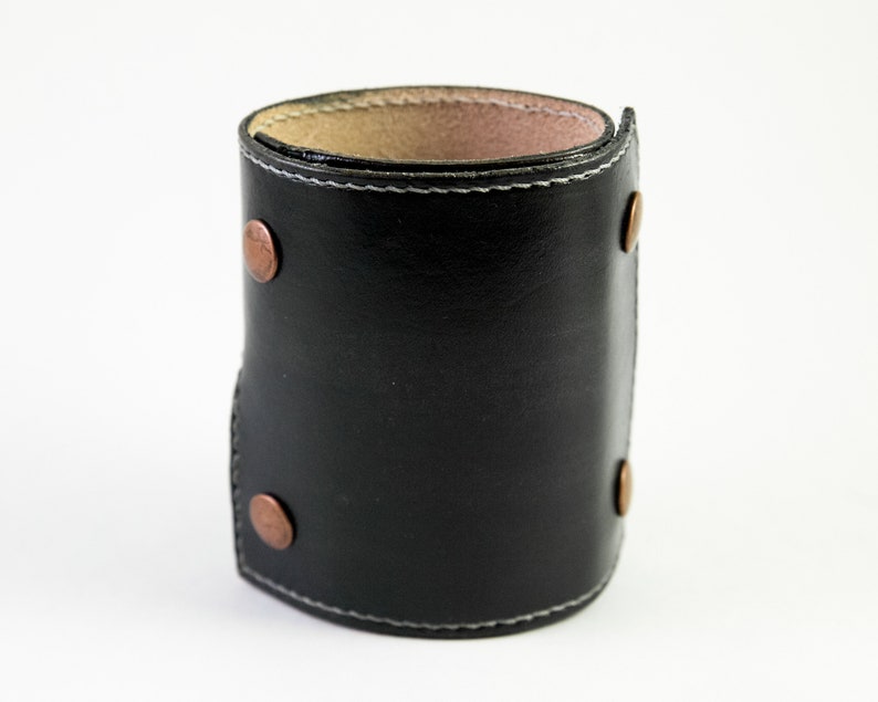 May include: A black leather cuff bracelet with copper rivets and white stitching. The cuff is folded over at the top and bottom.