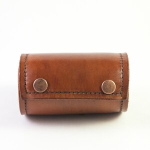 May include: A small, brown leather case with two brass-colored snap closures. The case is rectangular with rounded edges and appears to be hand-stitched. The leather has a natural, slightly distressed appearance.