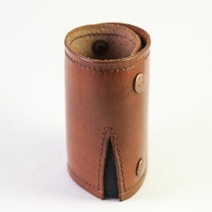 May include: A brown leather cuff bracelet with a rolled design. The bracelet features two copper-colored snap closures and a black fabric insert. The leather has visible stitching and a natural grain.