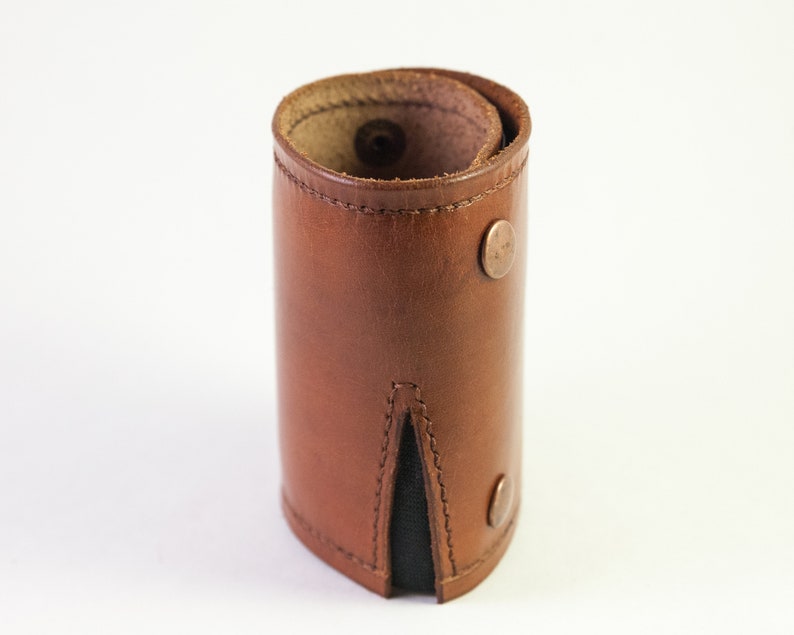 May include: A brown leather cuff bracelet with a rolled design. The bracelet features two copper-colored snap closures and a black fabric insert. The leather has visible stitching and a natural grain.