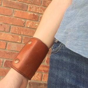 May include: A brown leather cuff bracelet with two copper-colored snap closures. The cuff is worn on the wrist. The background is a brick wall and the person is wearing a blue shirt and jeans.