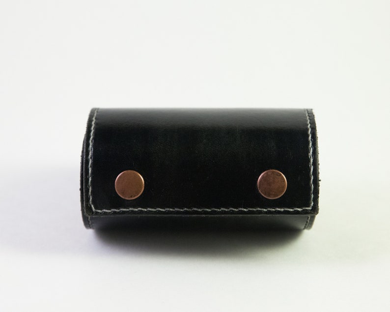 May include: A black leather roll-up case with two copper rivets. The case is closed and has a white stitch around the edges.