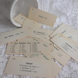 May include: A collection of vintage flashcards with German vocabulary words, scattered on a white surface. The cards are beige with black text, and some are partially visible. A white ceramic cup is in the background.