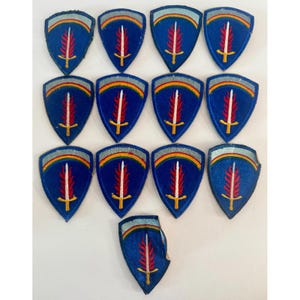 Puede incluir: Una colección de parches azules en forma de escudo. Cada parche presenta una espada blanca con un diseño rojo y amarillo, sobre un fondo azul. Un arco iris está encima de la espada.