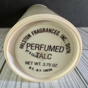 May include: A cylindrical container of Halston Fragrances, Inc. perfumed talc with a white base and black text. The label reads Halston Fragrances, Inc. Distr., Perfumed Talc, Net Wt. 3.75 oz., and N.Y., N.Y. 10036, with a small P107 code visible.