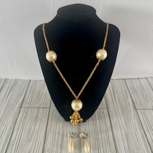 May include: A gold-toned necklace with three large, round, cream-colored faux pearls and a cluster of small gold beads. Matching earrings with cream-colored teardrop-shaped faux pearls are also displayed. The jewelry is presented on a black mannequin.