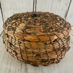 May include: A round, lidded basket crafted from coiled pine needles and natural fibers. The exterior features a textured, variegated brown and tan pattern with a dark, smooth, circular knob centered on the lid. The construction is dense and woven.