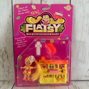 May include: A Flatsy doll set in a pink package. The package includes a small doll with long blonde hair, a yellow and orange outfit, and a miniature yellow house. The package reads "Flatsy, She's Flat and That's That!"